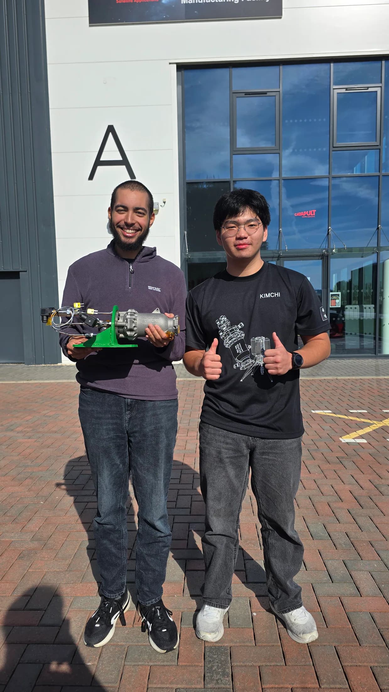 Two people outside a building entrance on test morning, one holding a small green engine mount.
