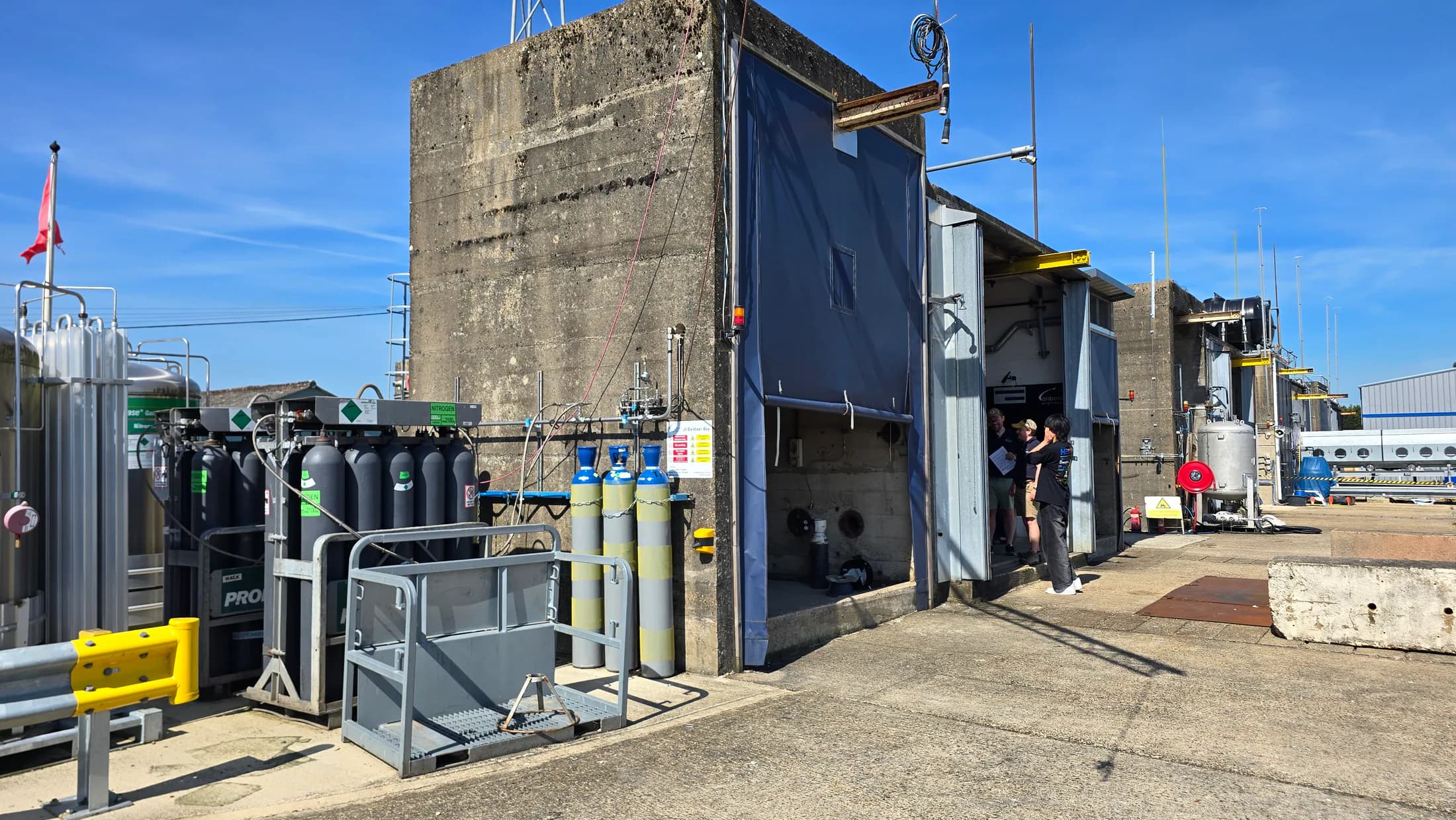 Exterior of the industrial test area with gas cylinders, concrete structures, and open test-bay doors.