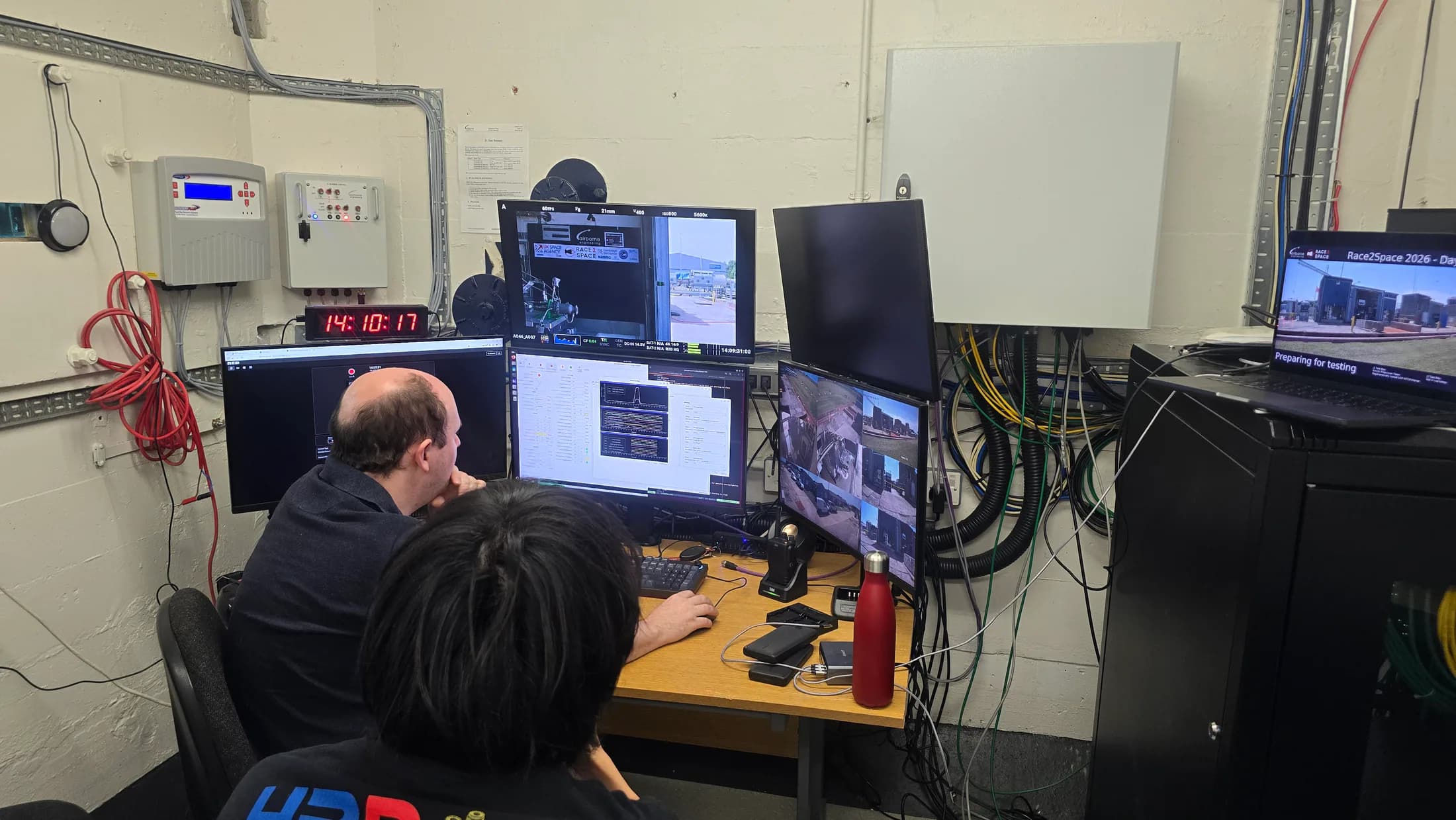Two people monitoring the test sequence at a workstation with multiple live camera and system feeds.