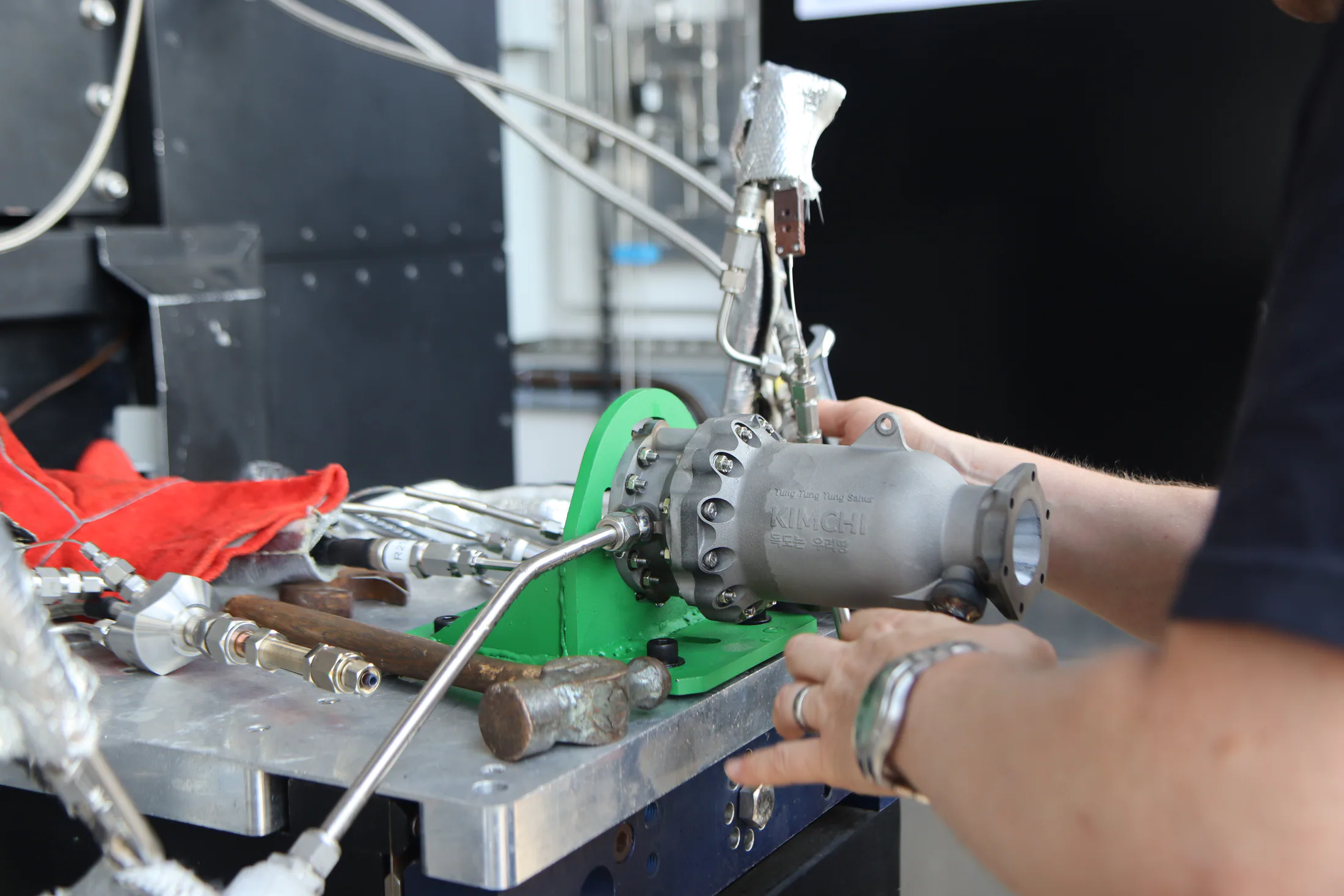 Hands adjusting the mounted cylindrical engine hardware with tubing and tools nearby.