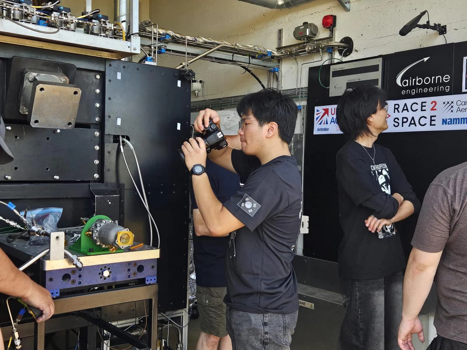 Team member documenting the engine setup while others stand nearby inside the facility.